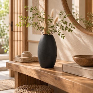 Könnte beinhalten: Eine mattschwarze, ovale Vase mit einer Ansammlung von grünen Blattzweigen und flauschigen, beigen Pampasgraswedeln. Die Vase hat eine glatte, abgerundete Silhouette und steht aufrecht als zentrales dekoratives Element im Raum.