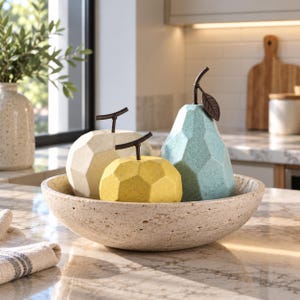 May include: A set of three geometric fruit sculptures in a beige, yellow, and light blue speckled finish, each topped with a dark brown metal stem and leaf, arranged inside a wide, shallow, textured beige stone bowl with a porous surface.