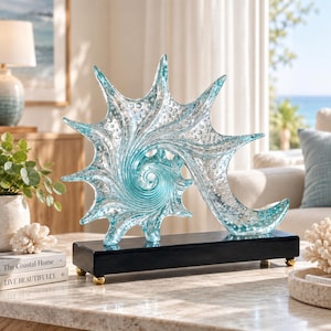 Peut inclure: Une sculpture décorative en forme de coquillage épineux, réalisée en verre transparent et bleu clair avec des bulles d'air internes. La pièce en verre est montée sur une base rectangulaire noire avec quatre petits pieds sphériques dorés.