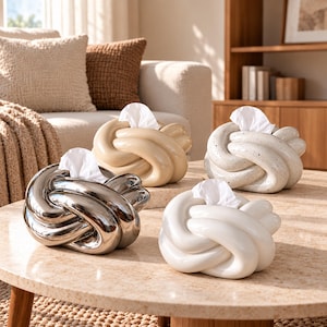 May include: Four knot-shaped tissue holders in different finishes: one metallic silver, one beige, one white with black speckles, and one solid white. Each holder features a central opening with a white tissue protruding from the top.