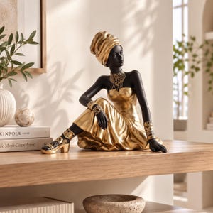 May include: A decorative figurine of a person with black skin wearing a gold strapless dress, a matching headwrap, a tiered necklace, and wide bracelets. The figure is seated, wearing gold heeled sandals with black accents, and features a smooth, matte finish on the skin and a metallic, reflective finish on the clothing and accessories.