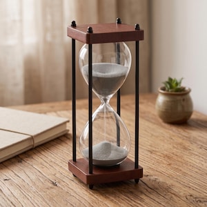 May include: An hourglass with a clear glass bulb containing fine gray sand, held within a frame featuring four thin black metal rods and square dark brown wooden top and bottom bases with small rounded feet at each corner of the base.