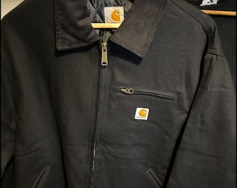 Könnte beinhalten: Eine schwarze Carhartt-Jacke mit Cordkragen, durchgehendem Frontreißverschluss und einer Reißverschlusstasche auf der Brust. Das Kleidungsstück verfügt über ein gestepptes Innenfutter und ein kleines, rechteckiges Carhartt-Markenlogo.