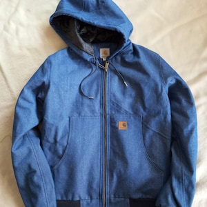 Puede incluir: Una chaqueta azul con capucha, cremallera frontal completa, mangas largas y puños y dobladillo acanalados en negro. La chaqueta cuenta con un forro acolchado negro en el interior de la capucha, un cordón y una etiqueta rectangular de Carhartt.