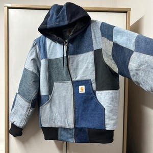 Könnte beinhalten: Eine Kapuzenjacke mit Reißverschluss aus einem Patchwork aus Denim-Stoff in verschiedenen Blau-, Hellblau- und Schwarztönen. Die Jacke verfügt über eine Kängurutasche auf der Vorderseite, einen schwarzen Rippsaum, schwarze Rippbündchen und ein Carhartt-Logo.