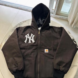 Peut inclure: Une veste à capuche zippée marron foncé avec un logo New York Yankees blanc sur la poitrine et une petite étiquette de marque Carhartt sur la poche avant. La veste présente des poignets côtelés noirs et une doublure intérieure matelassée noire.
