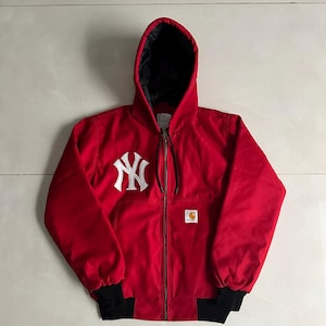 May include: A bright red hooded jacket featuring a white New York Yankees logo embroidered on the right chest and a Carhartt brand patch on the left pocket. The jacket has a black interior lining, a full-length front zipper, and black ribbed cuffs.