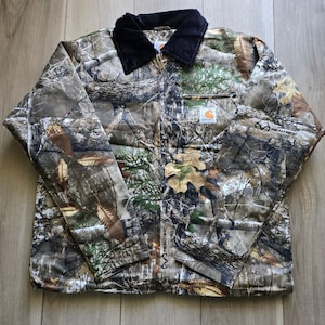 May include: A Carhartt jacket featuring a Realtree camouflage pattern with brown, green, and white forest elements. The jacket has a black corduroy collar, a full-length front zip, and a small Carhartt logo patch on the left chest pocket.