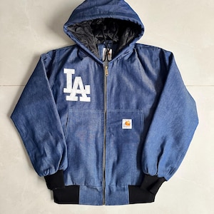 May include: A blue denim hooded jacket featuring a white Los Angeles Dodgers logo on the chest, a full-length front zip, black ribbed cuffs and hem, and a Carhartt brand patch on the front pocket with a black quilted interior lining.