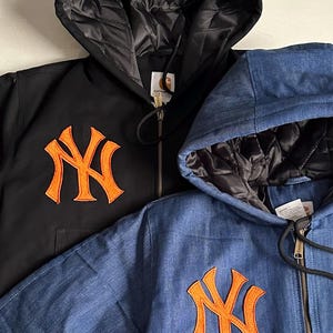 May include: Two zip-up hooded jackets, one in black and one in blue denim, each featuring a large orange embroidered New York Yankees logo on the chest. Both jackets have a black quilted lining inside the hood and a Carhartt brand label.