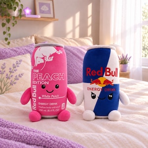 May include: Two plush toys shaped like energy drink cans. The left one is pink with white text reading The Peach Edition Red Bull, featuring a smiling face. The right one is blue and white with Red Bull branding and a smiling face with small arms and legs.