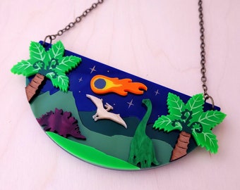 May include: A semi-circular pendant necklace featuring a prehistoric landscape with green palm trees, a purple stegosaurus, a green long-necked dinosaur, and a flying pterodactyl under a dark blue night sky with a bright orange and yellow meteor.