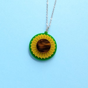 May include: A sunflower pendant necklace. The pendant features a yellow sunflower with a brown center set in a green circle. The necklace chain is silver-colored. The background is a light blue.