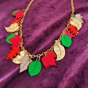 May include: A gold-colored chain necklace with multiple leaf charms in various colors, including red, green, gold, and gray. The leaves are arranged along the chain, creating a fall-themed accessory.