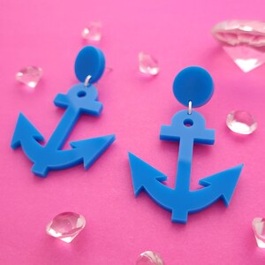 May include: A pair of bright blue anchor-shaped earrings featuring a circular stud top connected to a dangling anchor pendant. The earrings are made of a solid matte material and hang from silver-toned metal jump rings.