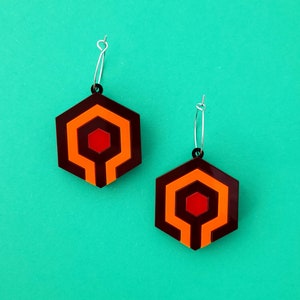 May include: A pair of hexagon-shaped earrings with a geometric design in shades of brown, orange, and red. The earrings have a silver hoop and a black accent at the top. The earrings are displayed against a teal background.