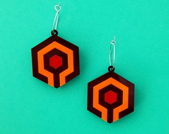 May include: A pair of hexagon-shaped earrings with a geometric design in shades of brown, orange, and red. The earrings have a silver hoop and a black accent at the top. The earrings are displayed against a teal background.
