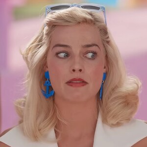 May include: A woman with blonde hair and blue eyes is wearing a white shirt and blue anchor earrings. She is looking to the side with a surprised expression.