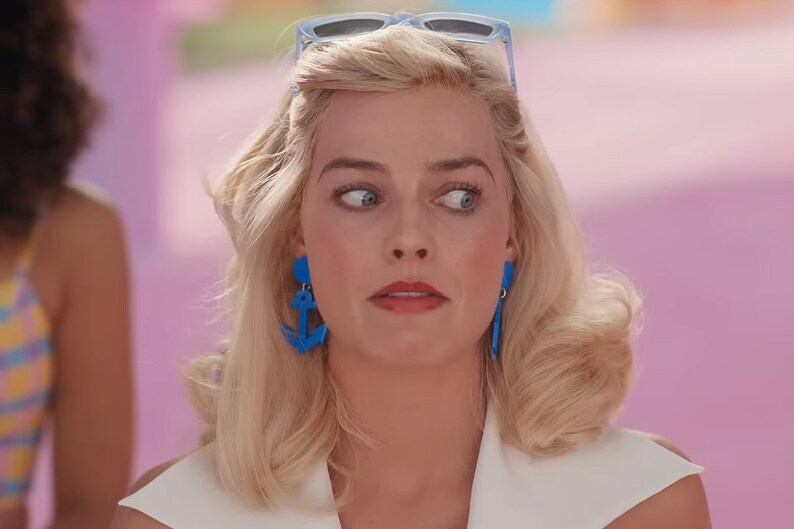 May include: A woman with blonde hair and blue eyes is wearing a white shirt and blue anchor earrings. She is looking to the side with a surprised expression.