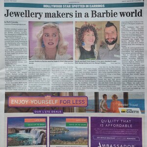 May include: A newspaper article features Margot Robbie wearing blue anchor earrings designed by Sugar & Vice. The article discusses the jewelry makers' surprise at seeing their work in the Barbie movie trailer.