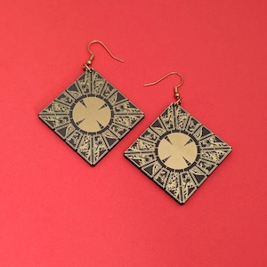 May include: A pair of square-shaped dangle earrings featuring a black and gold geometric pattern. Each earring has a central gold circle surrounded by intricate black and gold etched details in a diamond orientation with gold-toned metal hooks.
