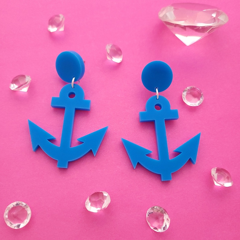 May include: A pair of bright blue anchor earrings with a circular stud top. The earrings are set against a vibrant pink background, with several clear, faceted crystals scattered around them. The earrings are a nautical-themed accessory.
