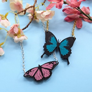 May include: Two butterfly necklaces. One necklace features a black butterfly with teal wings, and the other has a pink butterfly with black detailing. The necklaces hang from silver and gold chains, with pink and white flowers in the background.