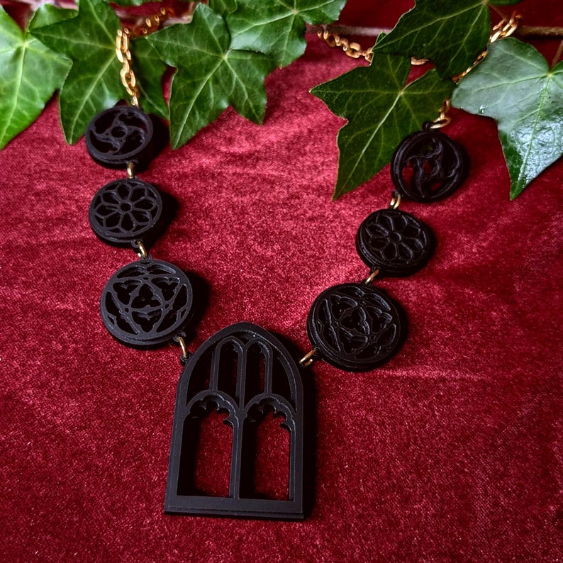 Puede incluir: Collar g&oacute;tico negro con cadena dorada. El collar presenta colgantes circulares con dise&ntilde;os intrincados y un colgante central en forma de ventana arqueada. El collar se exhibe sobre una superficie de terciopelo rojo con hiedra verde.