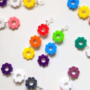 Könnte beinhalten: Hängeohrringe mit einer vertikalen Anordnung aus drei bunten Blumenanhängern aus Acryl. Die Blumen sind durch silberfarbene Metallringe verbunden und verfügen über einen Stecker mit einem transparenten Kunststoff-Ohrringverschluss.