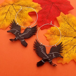 Puede incluir: Un par de pendientes de cuervo negro con aros plateados. Los cuervos están en vuelo con las alas extendidas.