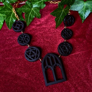 Puede incluir: Un collar g&oacute;tico negro con una cadena dorada. El collar presenta dise&ntilde;os de colgantes circulares y arqueados. Los colgantes tienen intrincados patrones calados. El collar se exhibe sobre una superficie de terciopelo rojo con hiedra verde.
