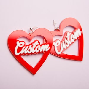 May include: A pair of red heart-shaped earrings featuring the word Custom in white script lettering across the center. The earrings are made of a smooth, glossy material and include silver-toned metal earring posts and clear plastic backings.