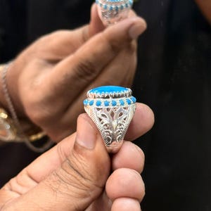 Peut inclure: Une bague en argent ornée d'une grande pierre ovale bleu vif sertie dans une monture dentelée. L'anneau est décoré d'une rangée de petites pierres rondes bleues et de motifs métalliques ajourés de style filigrane sur les côtés.