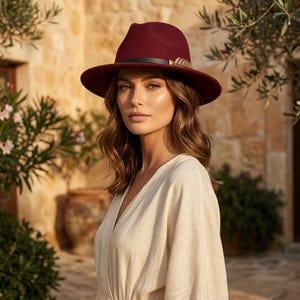 May include: A deep burgundy felt fedora hat featuring a black leather band accented with a metallic gold-toned feather detail. The hat has a structured crown and a wide brim, providing a classic silhouette that complements the beige linen garment.