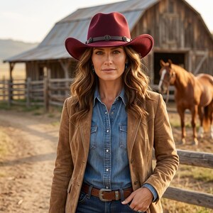 Puede incluir: Un sombrero de vaquero de fieltro rojo oscuro con una banda de cuero marrón oscuro que presenta pequeños detalles metálicos circulares. Se lleva con una camisa vaquera azul, una americana de ante beige y un cinturón de cuero marrón con hebilla plateada.