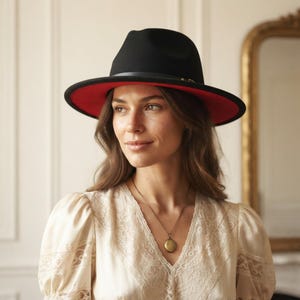 May include: A black fedora hat featuring a wide brim with a vibrant red underside and a thin black band around the crown. The hat is worn with a cream-coloured blouse that has lace detailing on the sleeves and a gold locket necklace.