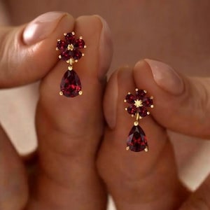 May include: A pair of gold earrings featuring a flower-shaped cluster of deep red garnet stones at the top, with a single pear-shaped garnet drop suspended below each flower. The metal setting is a warm yellow gold tone with a polished finish.