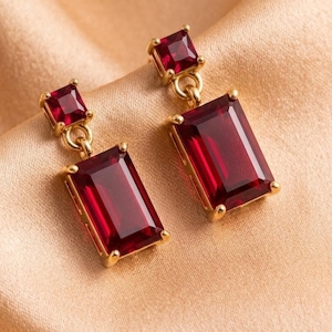 May include: A pair of gold drop earrings featuring a small square-cut deep red gemstone at the top, connected by a jump ring to a larger rectangular-cut deep red gemstone. Each stone is secured in a four-prong gold setting with a polished finish.