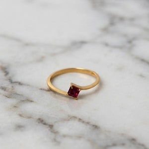 Op de afbeelding: Een dunne gouden ring met een enkele, kleine, vierkant geslepen dieprode edelsteen in het midden. De ringband is slank en glad, waarbij het metaal iets taps toeloopt naar de zetting van de levendige, donkerrode vierkante steen.