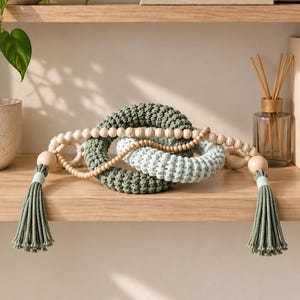 May include: A decorative garland featuring two interlocking crocheted rings in sage green and light blue, strung with natural wooden beads. The ends of the bead strand are finished with sage green cotton tassels secured by light-colored bands.