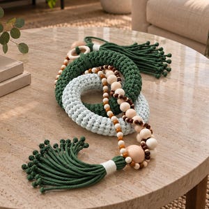 May include: A decorative chain consisting of two interlocking crocheted rings in dark green and light blue, accented with a long strand of natural wooden beads in varying sizes and shades, finished with dark green knotted tassels at each end.