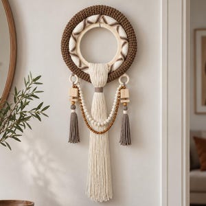Könnte beinhalten: Ein kreisförmiger Wandbehang mit einem dicken, braun gehäkelten Außenring und einem cremefarben umwickelten Innenreifen. Er enthält eine lange, cremefarbene Quaste in der Mitte, zwei kleinere graue Quasten, Holzperlen und quadratische Holzblöcke.