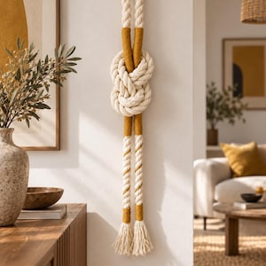 Pode incluir: Um adorno de parede composto por uma corda grossa trançada na cor creme, atada num grande nó decorativo. A corda é acentuada com secções de fio amarelo-mostarda enroladas firmemente, terminando em duas borlas desfiadas na parte inferior.