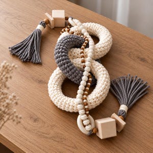 May include: A decorative chain featuring two interlocking crocheted rings in cream and dark gray, accented with a strand of white and brown wooden beads, two light wood cubes, and two dark gray rope tassels at the ends of the assembly.