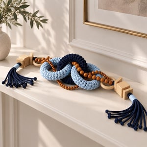 May include: A decorative chain featuring three interlocking rings covered in navy blue and light blue crochet yarn, accented with natural wooden beads, square wooden blocks, and long navy blue tassels at both ends of the horizontal arrangement.