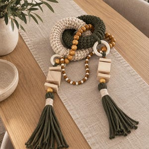 May include: A decorative garland featuring two macrame rings in cream and dark green, connected by strings of polished wooden beads. The piece includes square wooden blocks and two long dark green tassels, creating a textured, multi-layered hanging ornament with natural tones and varied geometric shapes.
