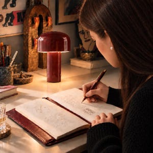 May include: A glossy dark red mushroom-shaped table lamp with a cylindrical base and a wide, rounded shade. The lamp emits a warm, soft glow, illuminating an open notebook with cream-coloured pages and a matching dark red fountain pen.