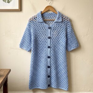Peut inclure: Une chemise en crochet bleu clair dotée d'un col, de manches courtes et d'une fermeture boutonnée sur le devant. Le vêtement est réalisé avec un motif géométrique ajouré et comporte six boutons bleu foncé alignés sur le milieu.