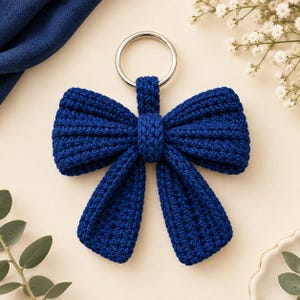 May include: A dark blue crocheted bow keychain featuring a silver-toned metal ring. The bow is constructed with a textured, knotted yarn pattern, creating a structured shape with two loops and two hanging tails attached to the circular metal ring.