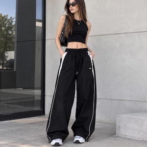 Peut inclure: Un haut court noir sans manches associé à un pantalon de survêtement noir ample doté de bandes latérales blanches, d'une taille à cordon de serrage et de stoppeurs réglables à l'ourlet. Le pantalon présente un petit logo blanc sur la cuisse.